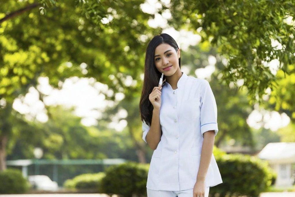 Femme en uniforme de soins, souriant et tenant un stylo, avec des arbres en arrière-plan.