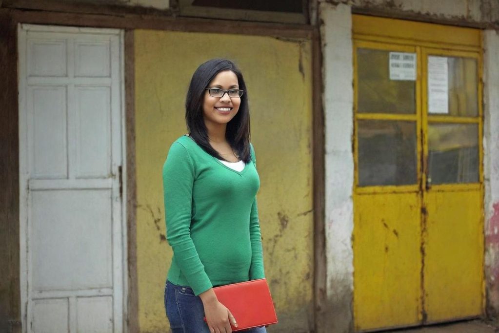 Femme souriante en pull vert, tenant un carnet, devant un mur jaune et des portes.
