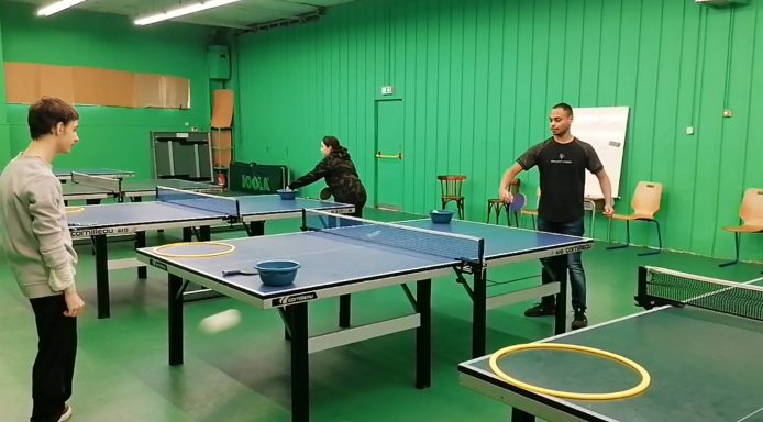 Deux personnes jouent au ping-pong dans une salle avec des tables et un mur vert.