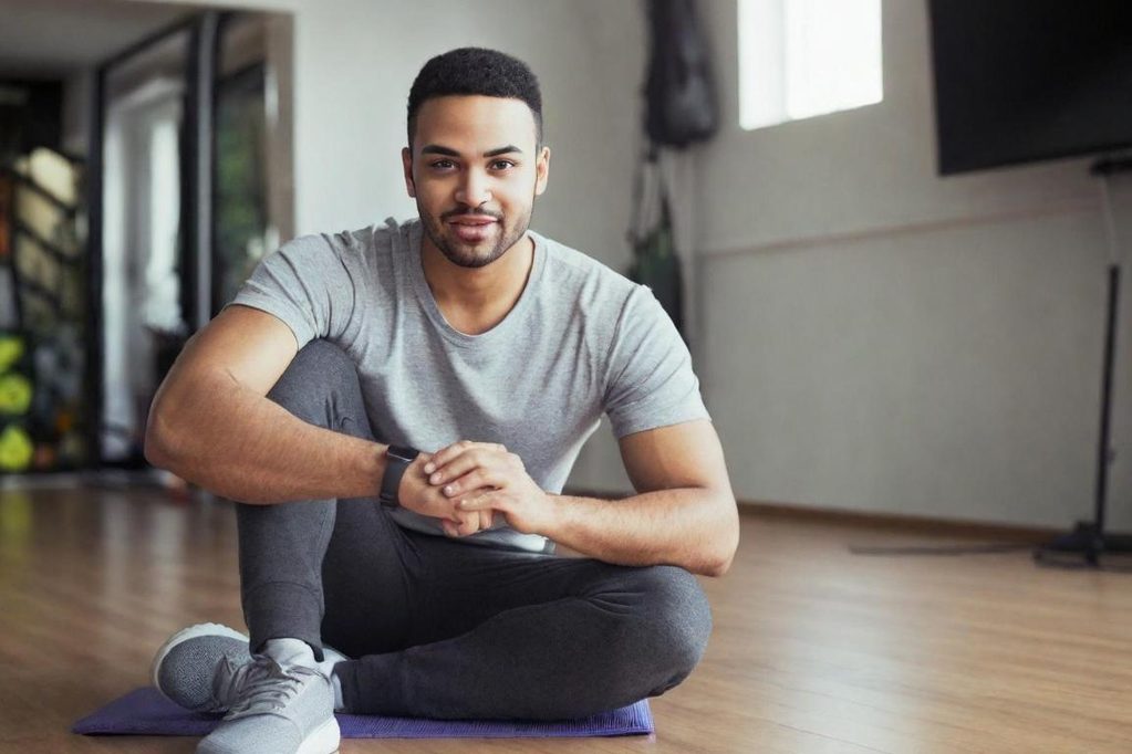 Un homme souriant assis sur un tapis de yoga dans une salle de sport.