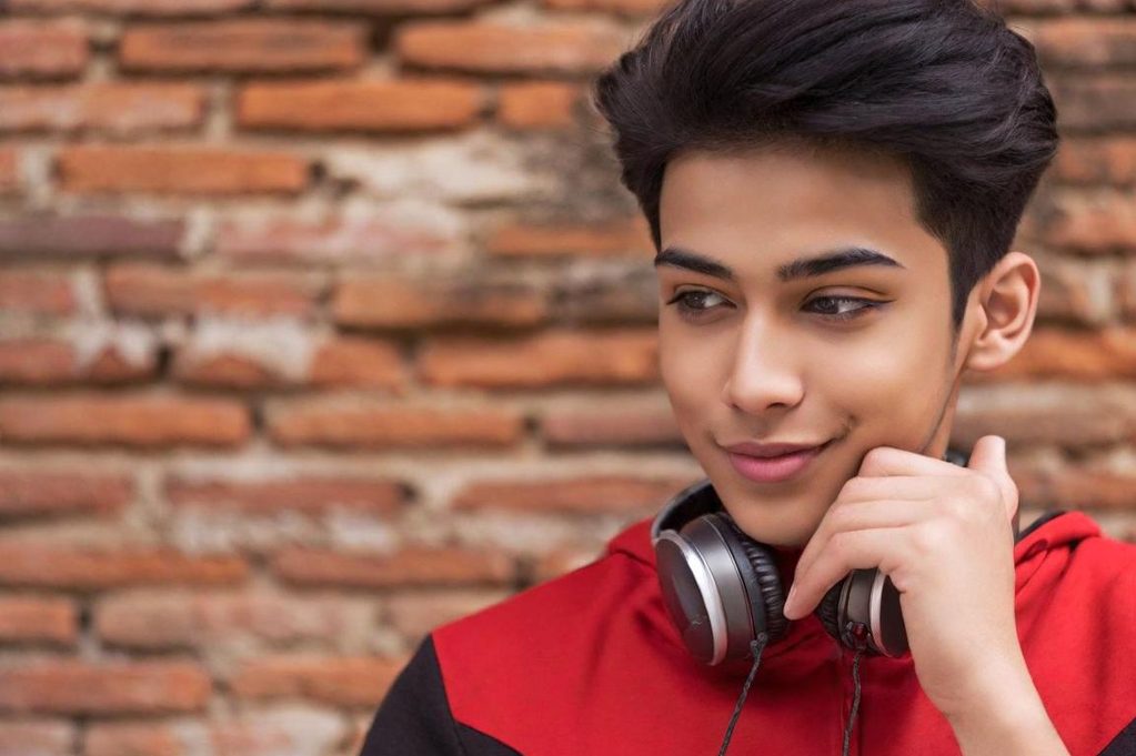 Jeune homme souriant portant des écouteurs, devant un mur en briques.