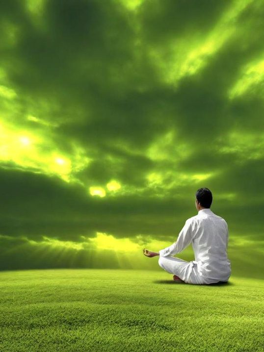 Un homme en méditation, assis sur une herbe verte sous un ciel nuageux et lumineux.