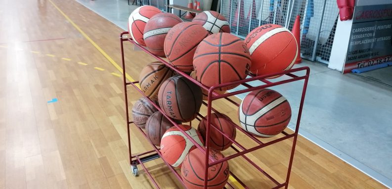 Un chariot rempli de ballons de basket orange et rouge sur un parquet de gymnase.